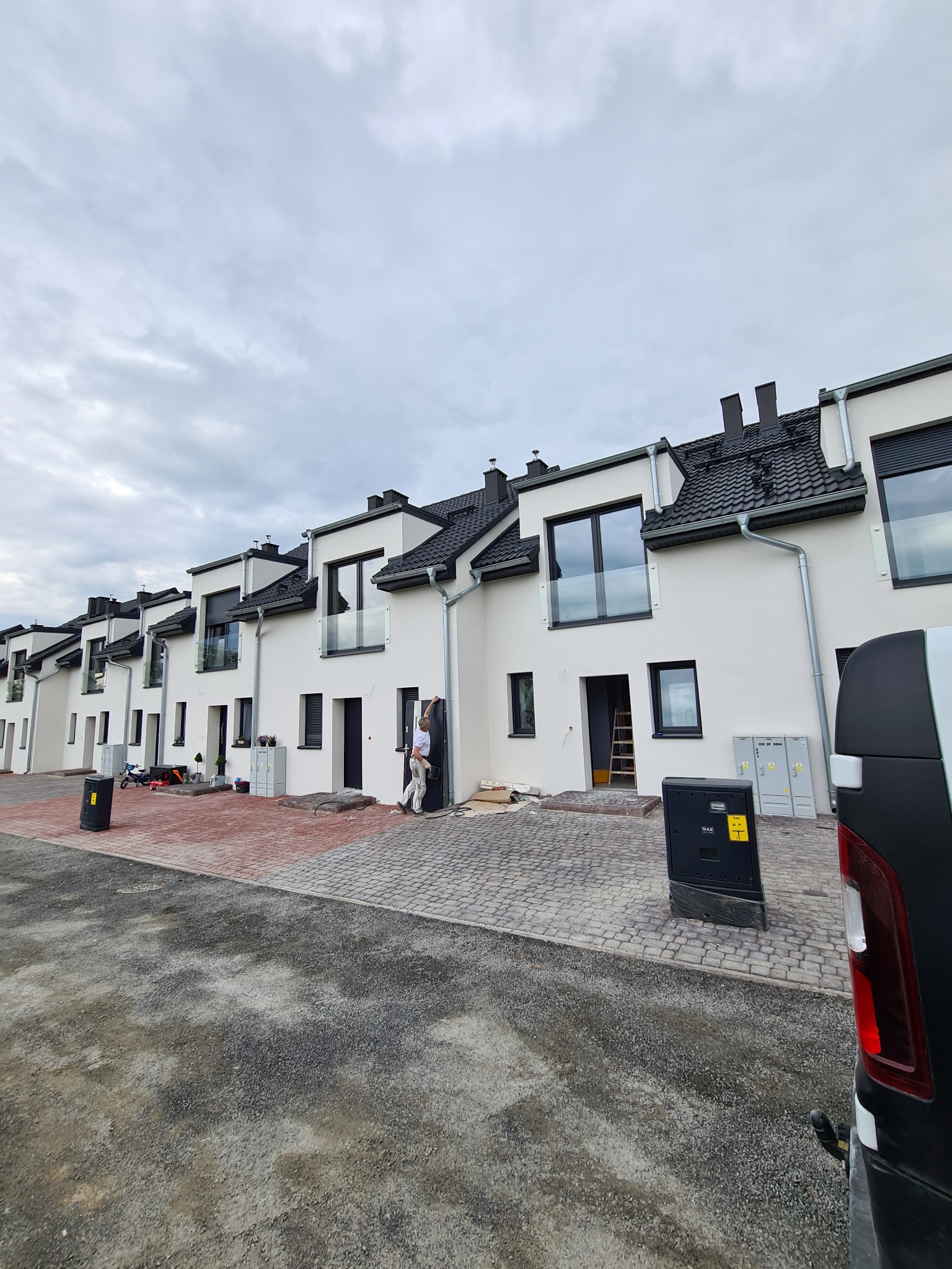 Szereg nowo wybudowanych, białych domów szeregowych z czarnymi dachami i oknami, z pracownikiem montującym szklaną balustradę balkonową. Widoczny fragment chodnika z kostki brukowej i samochód...