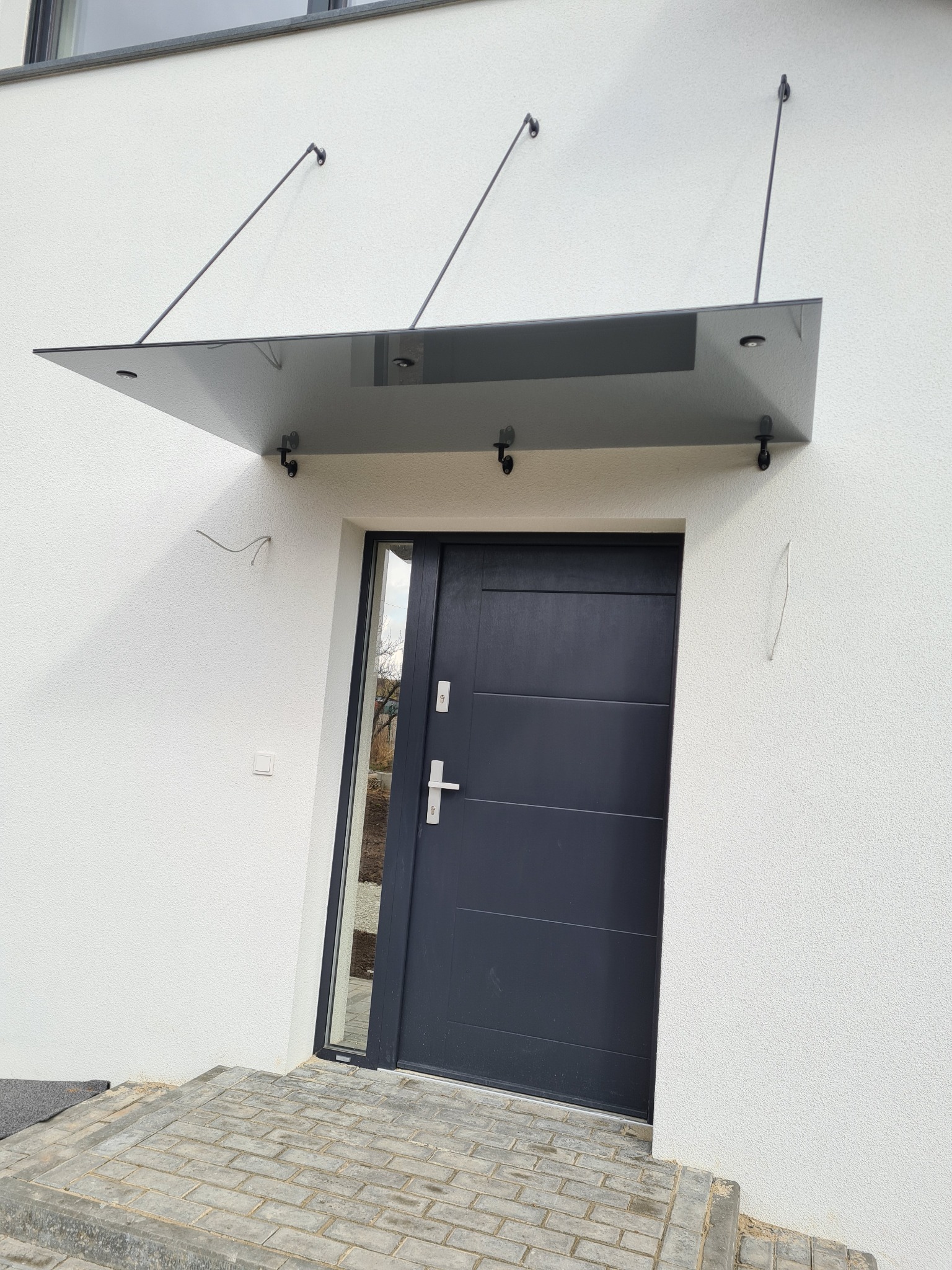 Szklany daszek nad wejściem do domu, wsparty na metalowych wspornikach, zamontowany nad ciemnymi drzwiami wejściowymi i kostką brukową.