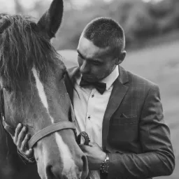 Czarno-białe zdjęcie mężczyzny w garniturze z muchą, trzymającego za uzdę brązowego konia z białą strzałką na czole.