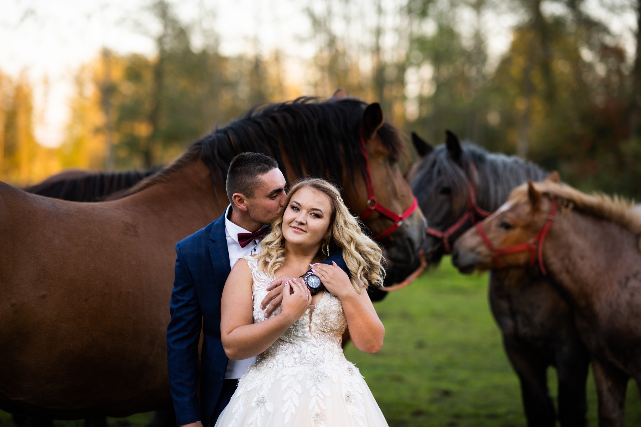 Para młoda w objęciach na tle koni na łące, panna młoda w sukni ślubnej z koronkowymi aplikacjami, pan młody w granatowym garniturze w kratę, pocałunek w policzek.