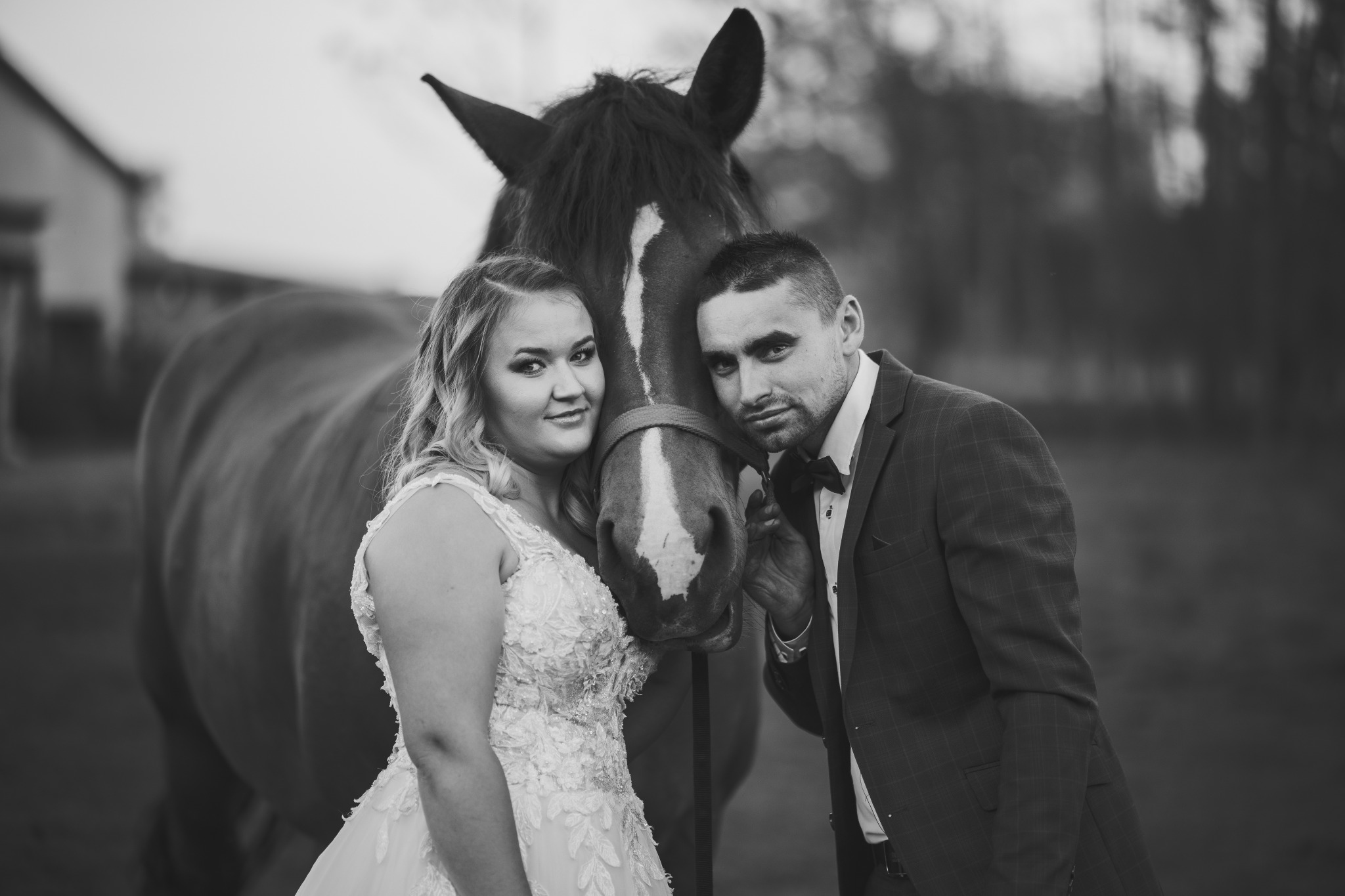 Czarno-białe zdjęcie pary młodej w strojach ślubnych, pozującej z koniem z białą strzałką na pysku.