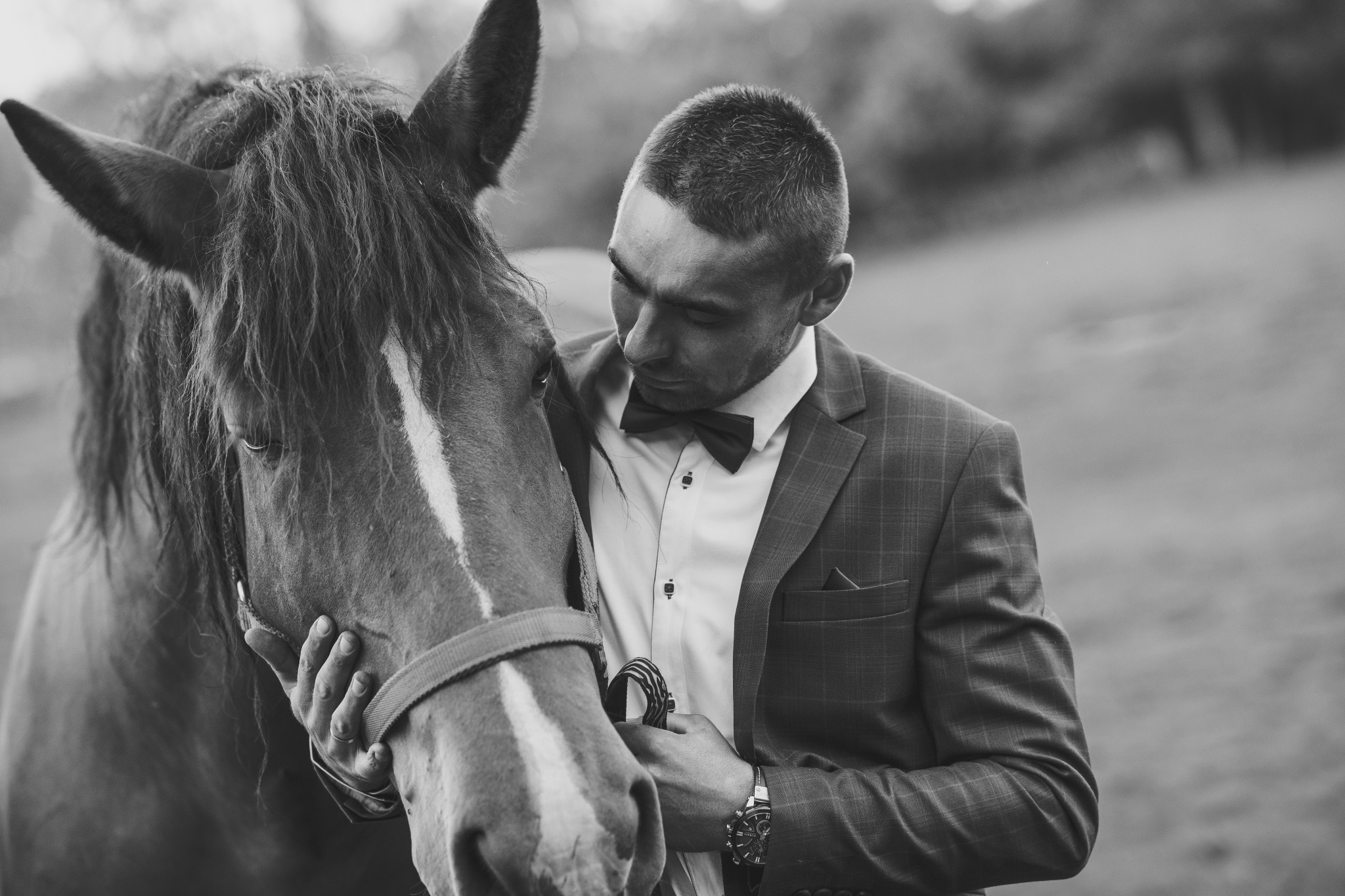 Czarno-białe zdjęcie mężczyzny w garniturze z muchą, trzymającego za uzdę brązowego konia z białą strzałką na czole.