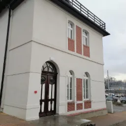 Dwupiętrowy budynek z białą elewacją, ciemnymi drewnianymi drzwiami z łukowym nadprożem i ceglanymi wstawkami wokół okien, widok z zewnątrz na parking w tle.