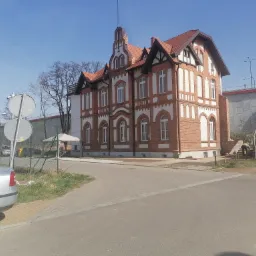 Odrestaurowany, dwupiętrowy dom murowany z czerwonej cegły z białymi zdobieniami, czerwoną dachówką i elementami szachulcowymi, widoczny z ulicy w słoneczny dzień.