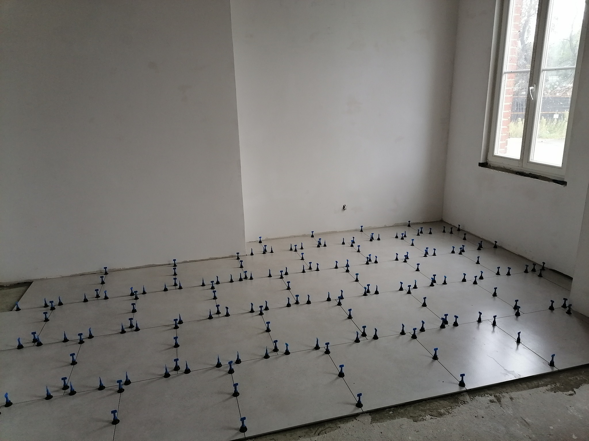 Układanie jasnych płytek podłogowych w nowym pomieszczeniu z użyciem systemu poziomowania, widoczne niezagruntowane ściany i okno.