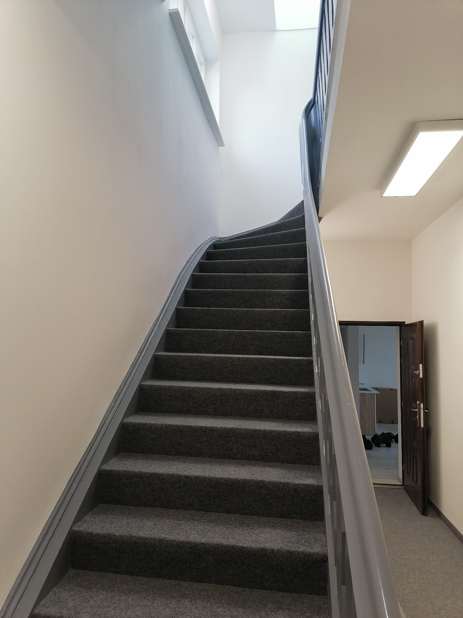 Wewnętrzne schody z szarą wykładziną i szarą balustradą prowadzące na wyższe piętro, widoczne drzwi do pomieszczenia z wyposażeniem.