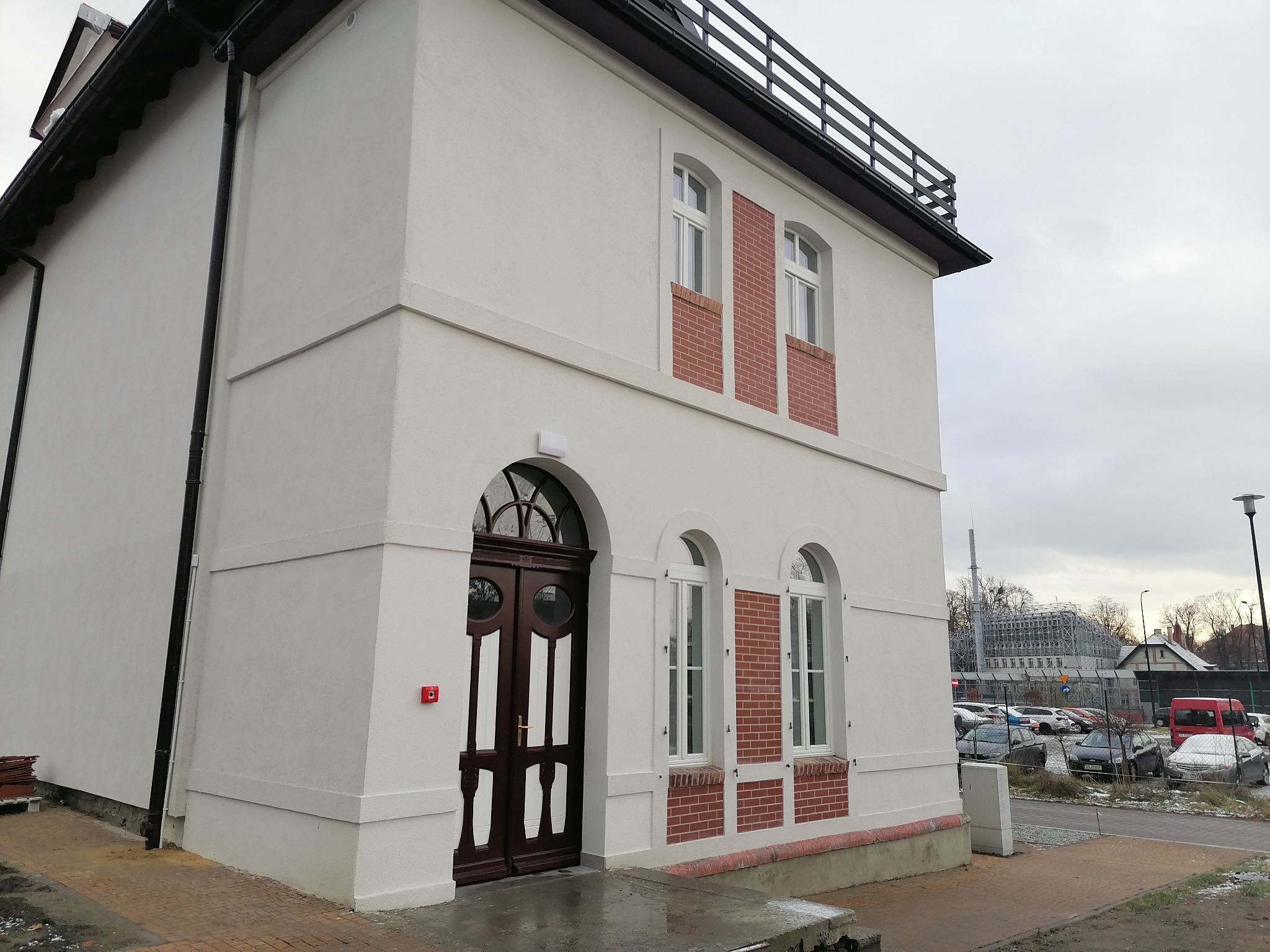 Dwupiętrowy budynek z białą elewacją, ciemnymi drewnianymi drzwiami z łukowym nadprożem i ceglanymi wstawkami wokół okien, widok z zewnątrz na parking w tle.