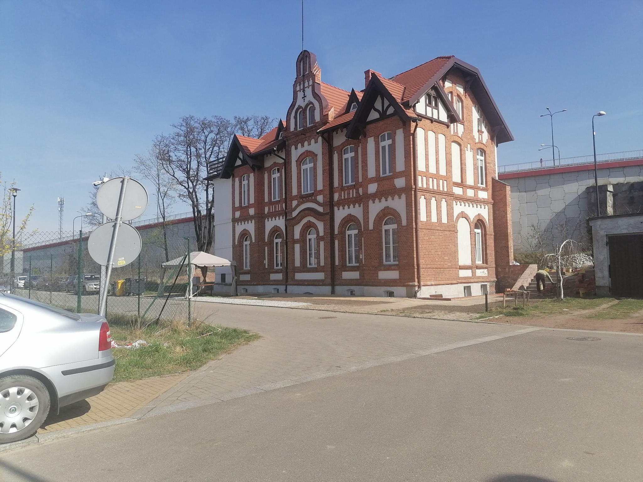 Odrestaurowany, dwupiętrowy dom murowany z czerwonej cegły z białymi zdobieniami, czerwoną dachówką i elementami szachulcowymi, widoczny z ulicy w słoneczny dzień.