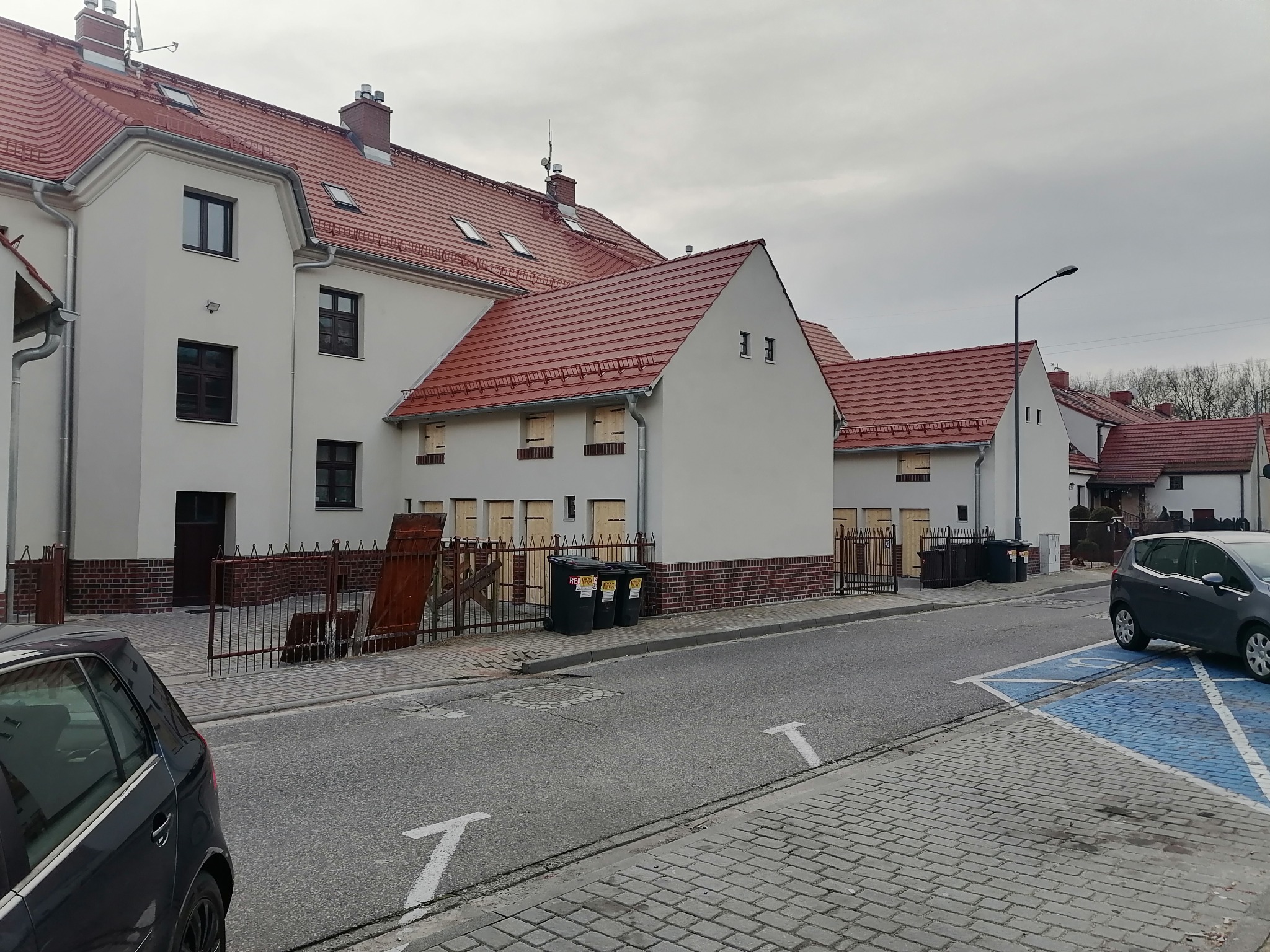 Nowe, białe budynki mieszkalne z czerwoną dachówką, drewnianymi okiennicami i ceglanymi detalami, widoczne z poziomu ulicy z zaparkowanymi samochodami i oznaczeniami miejsc parkingowych.