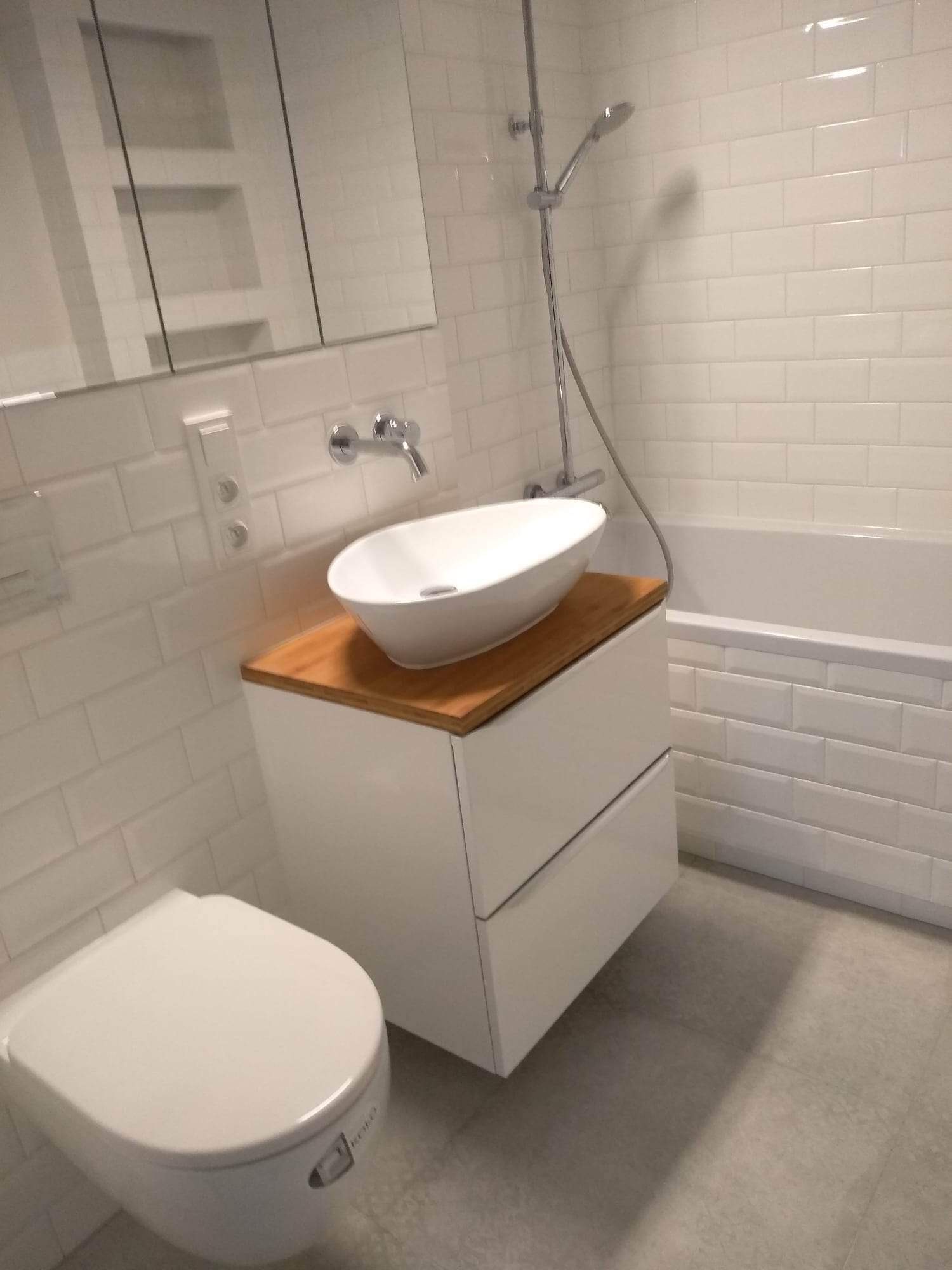 Nowoczesna łazienka z białą armaturą, owalną umywalką na drewnianym blacie, białą wanną i toaletą podwieszaną, wykończona białymi, prostokątnymi płytkami.