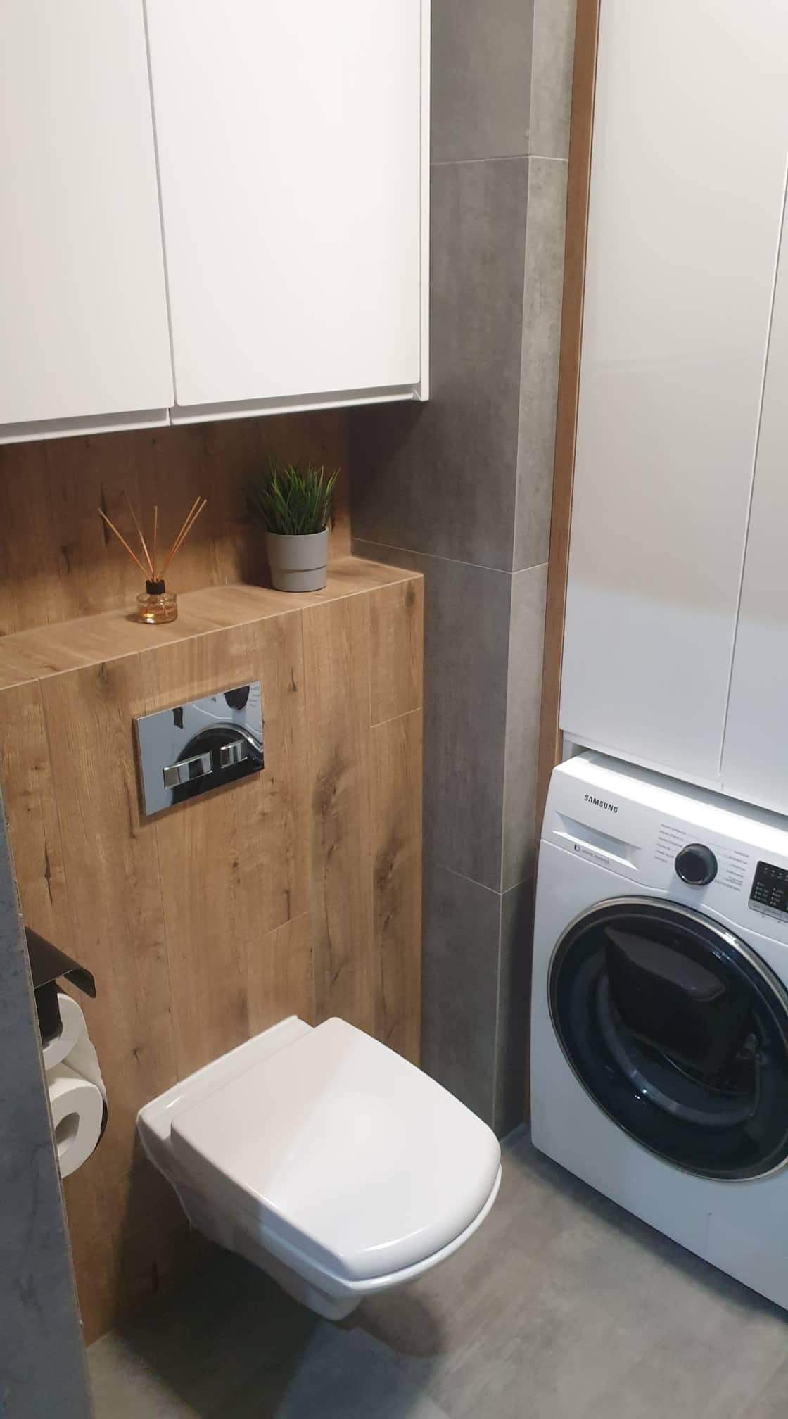 Nowoczesna łazienka z podwieszaną toaletą, zabudową z drewnopodobnych paneli, szarą płytką i pralką Samsung.