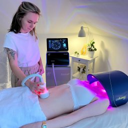 Salon Urody Skelnik Agnieszka Face & Body - Kobieta wykonuje zabieg na brzuchu pacjentki za pomocą urządzenia Celluma Pro w jasnym gabinecie, oświetlonym lampą. Widoczny sprzęt kosmetyczny.