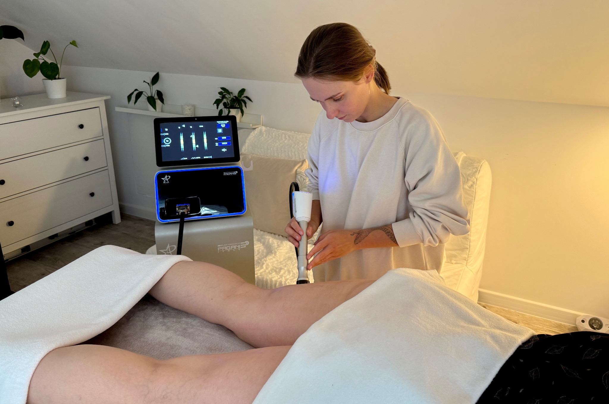 Kobieta wykonuje zabieg na udzie pacjenta za pomocą nowoczesnego urządzenia w jasnym gabinecie. Widoczny panel sterowania urządzenia.