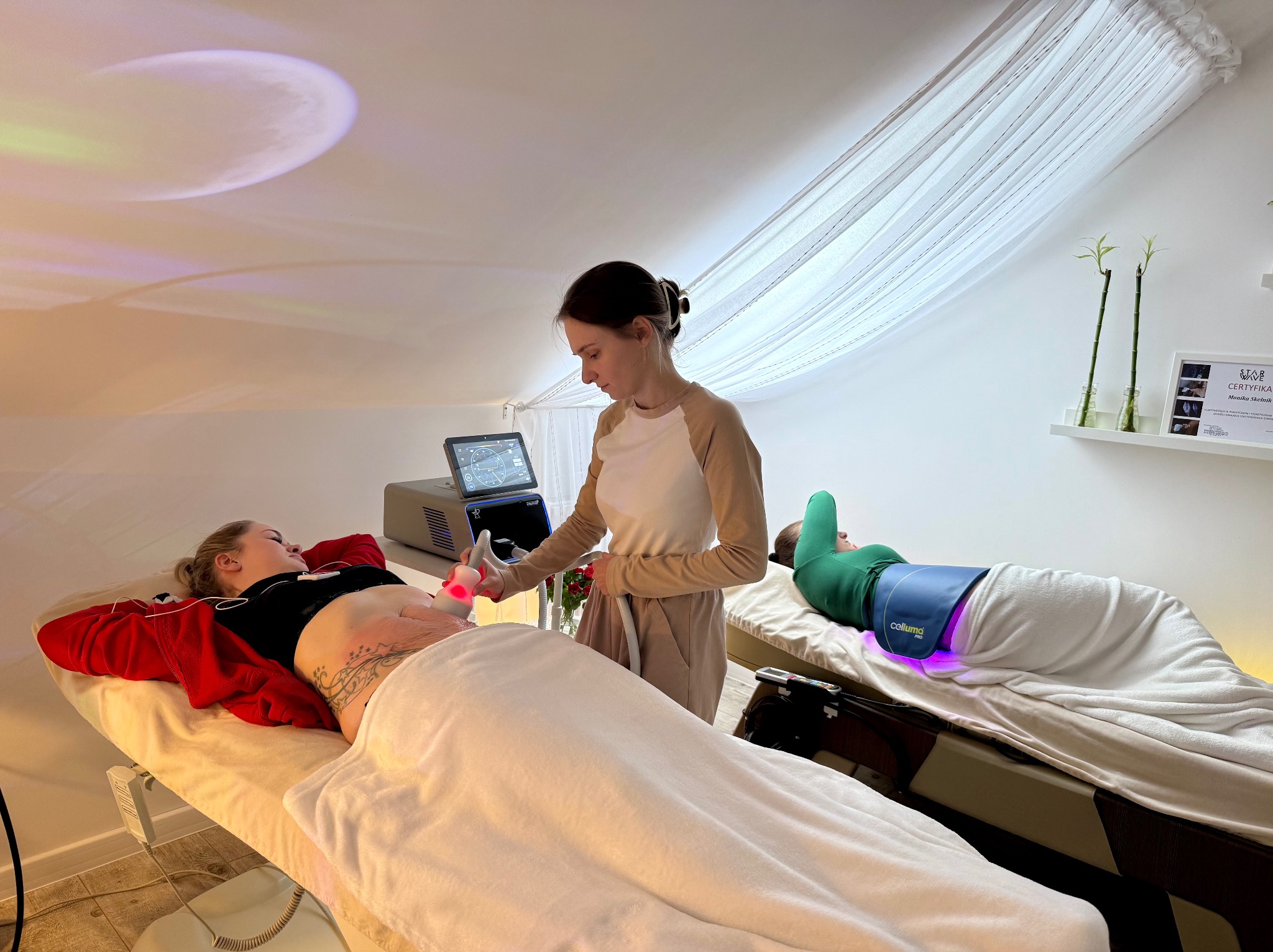 Kobieta wykonuje zabieg urządzeniem na brzuchu pacjentki leżącej na leżance w gabinecie. W tle druga pacjentka poddaje się zabiegowi z użyciem pasa.