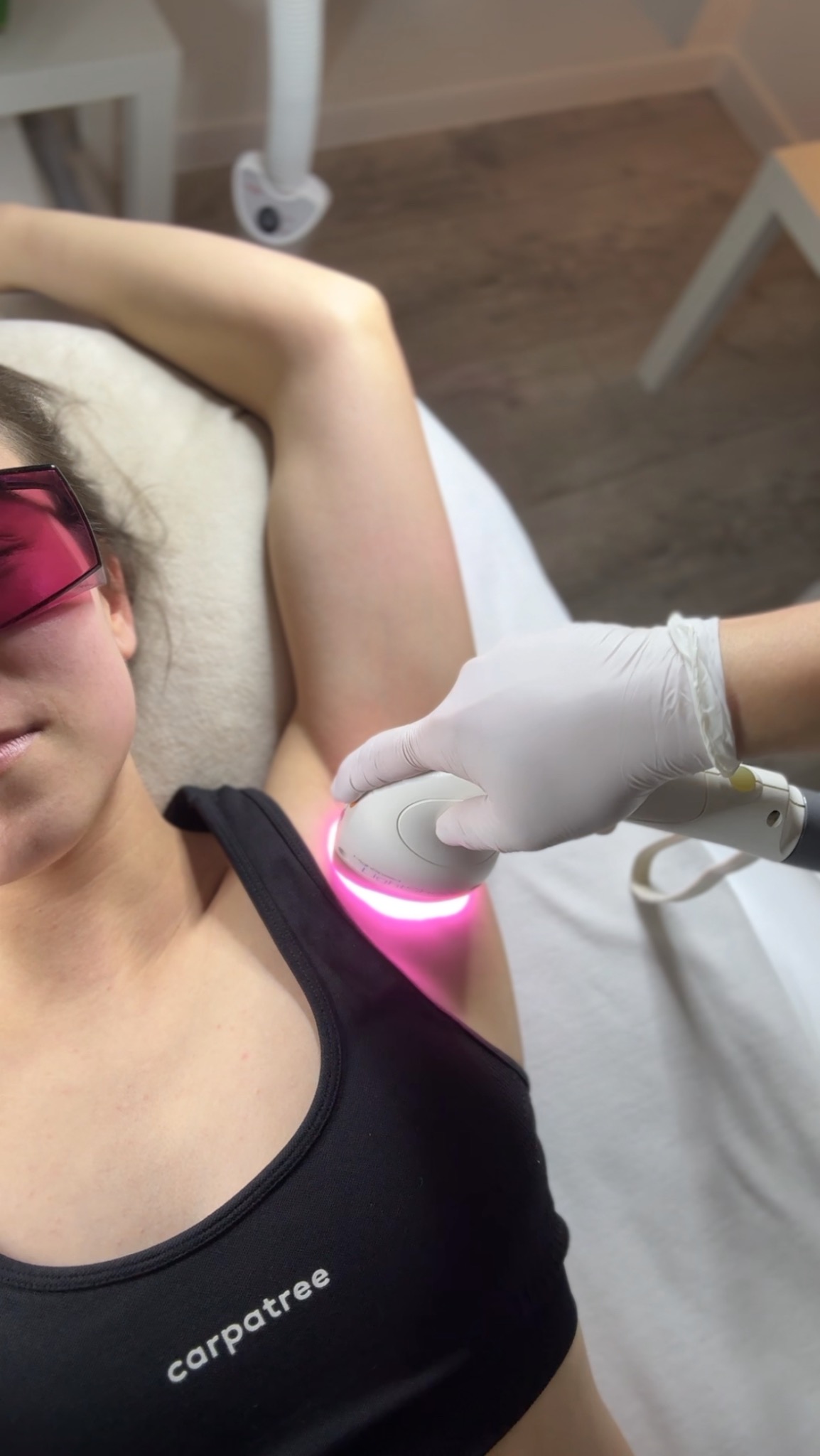 Osoba w okularach ochronnych poddawana zabiegowi depilacji pach pod pachami, wykonywanemu przez specjalistę w rękawicach ochronnych przy użyciu urządzenia laserowego.