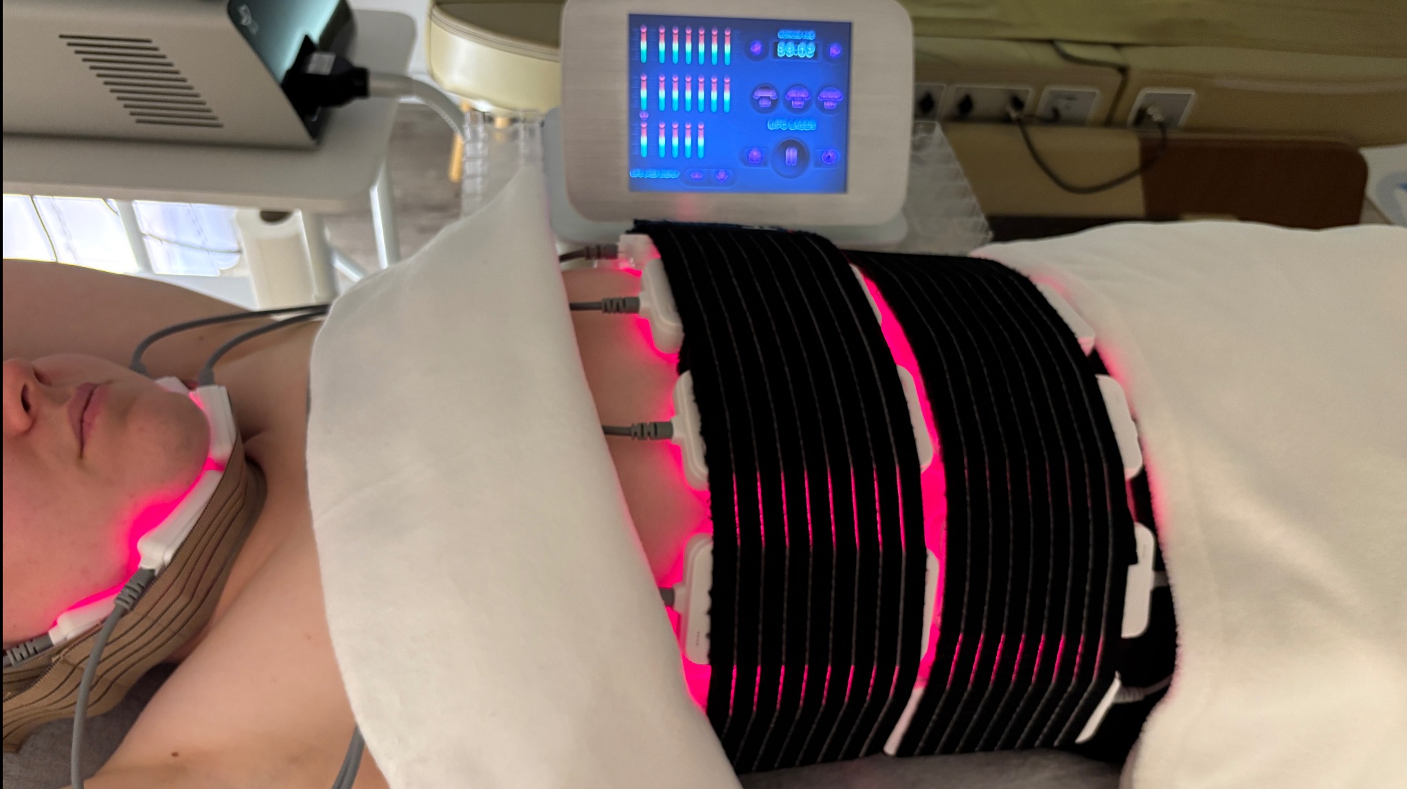 Osoba poddawana zabiegowi modelowania sylwetki z użyciem urządzenia emitującego czerwone światło. Widoczne panele urządzenia i fragment twarzy klienta.