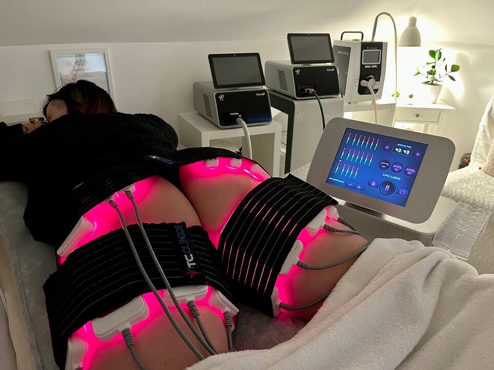 Klientka podczas zabiegu lipo laser na uda w salonie. Widoczne czarne pasy z czerwonym światłem LED i aparatura kontrolna z ekranem.
