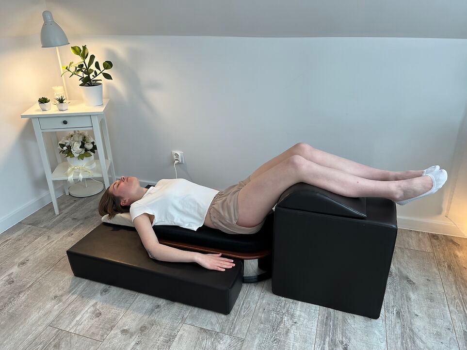 Kobieta leży na specjalistycznym fotelu do masażu, z nogami uniesionymi na podwyższeniu. W tle stolik nocny z lampą i roślinami. Jasne wnętrze.