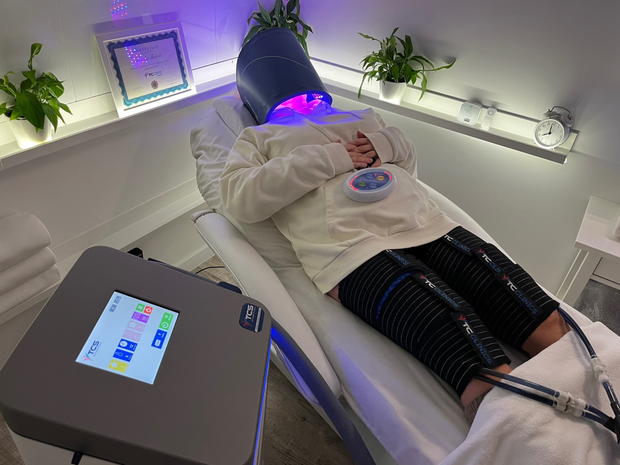 Klientka w trakcie zabiegu z użyciem specjalistycznego sprzętu w jasnym gabinecie, widoczny certyfikat i rośliny doniczkowe.