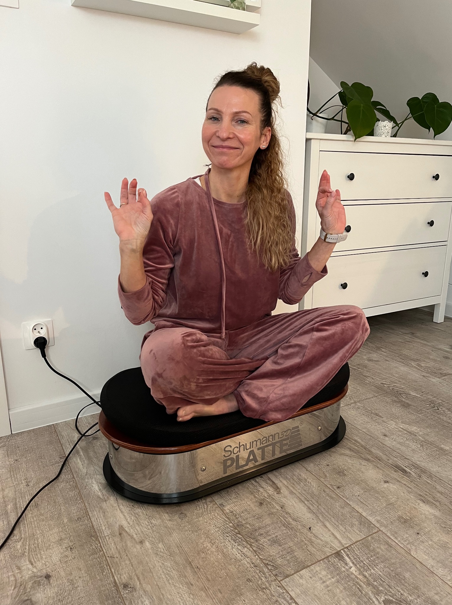 Kobieta w welurowym dresie siedzi po turecku na platformie wibracyjnej Schumann 3D Platte, unosząc dłonie. W tle biała komoda i roślina doniczkowa.