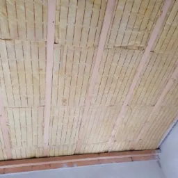 Izolacja z wełny mineralnej z perforacją, zamontowana między drewnianymi belkami stropu poddasza, widoczne szczeliny między płytami izolacyjnymi.