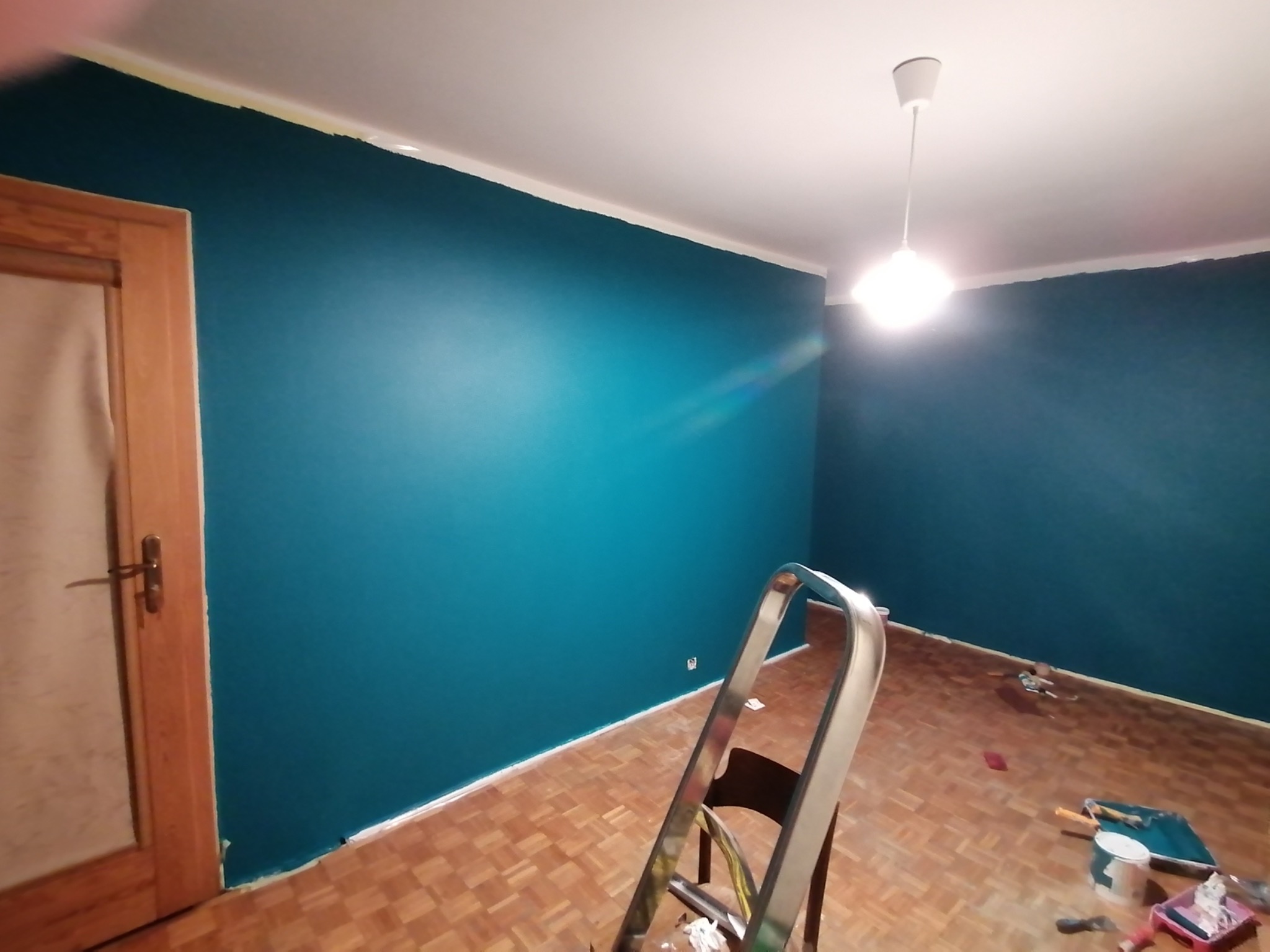 Świeżo pomalowane na turkusowo ściany w pokoju, widoczna drabina, podłoga parkietowa, narzędzia malarskie i taśma malarska przy suficie.