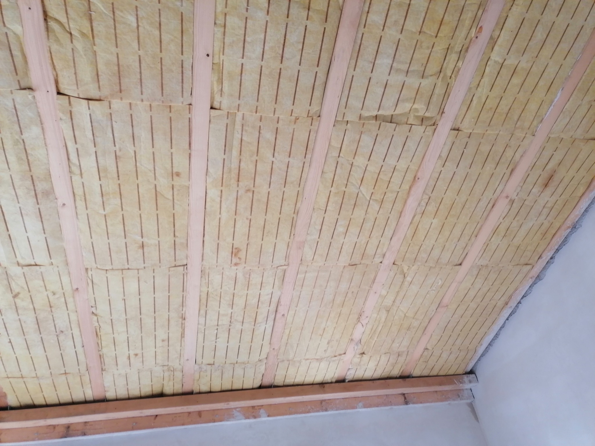 Izolacja z wełny mineralnej z perforacją, zamontowana między drewnianymi belkami stropu poddasza, widoczne szczeliny między płytami izolacyjnymi.