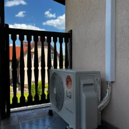 Zewnętrzna jednostka klimatyzacji LG Dual Inverter zamontowana na balkonie, widok przez czarną, drewnianą balustradę na dom z czerwoną dachówką i błękitne niebo z chmurami.