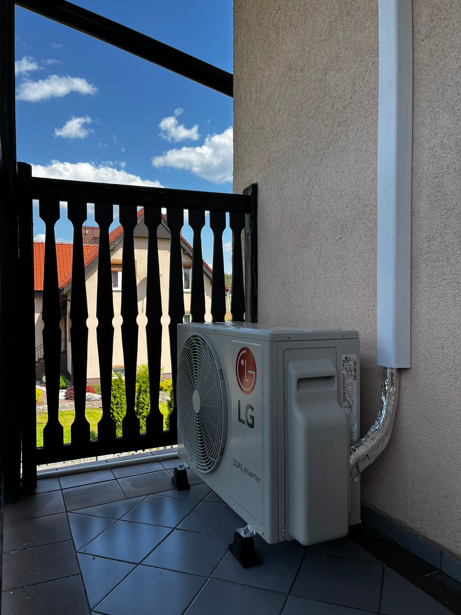 Zewnętrzna jednostka klimatyzacji LG Dual Inverter zamontowana na balkonie, widok przez czarną, drewnianą balustradę na dom z czerwoną dachówką i błękitne niebo z chmurami.