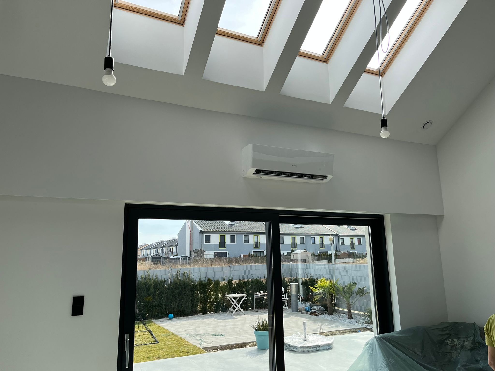 Klimatyzator GREE zamontowany na ścianie w nowoczesnym salonie z oknami dachowymi i widokiem na ogród z palmami.