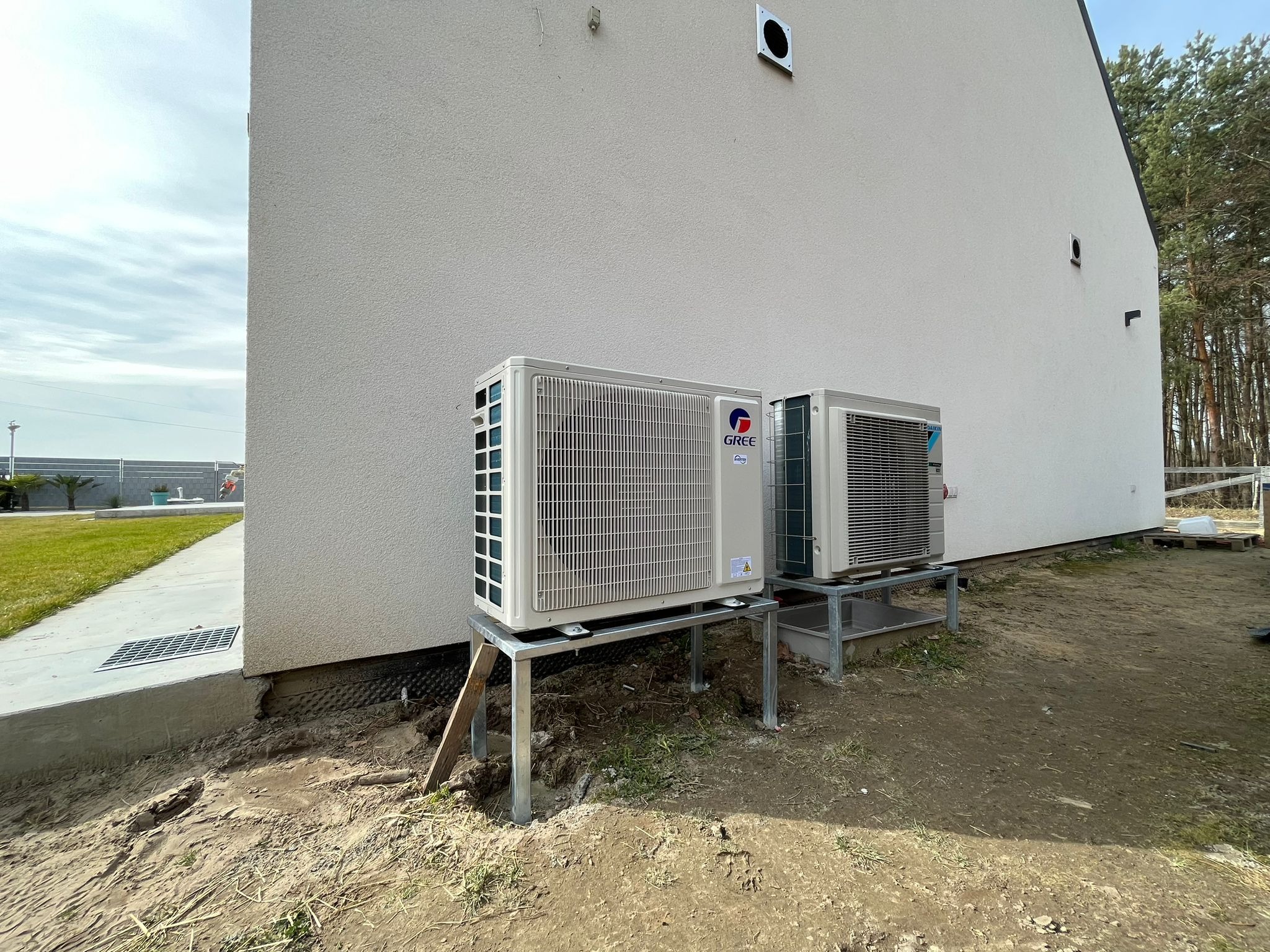 Dwa zewnętrzne agregaty klimatyzacji GREE i DAIKIN zamontowane na metalowych stelażach przy ścianie budynku z jasną elewacją, widoczne elementy instalacji i teren wokół w trakcie prac.