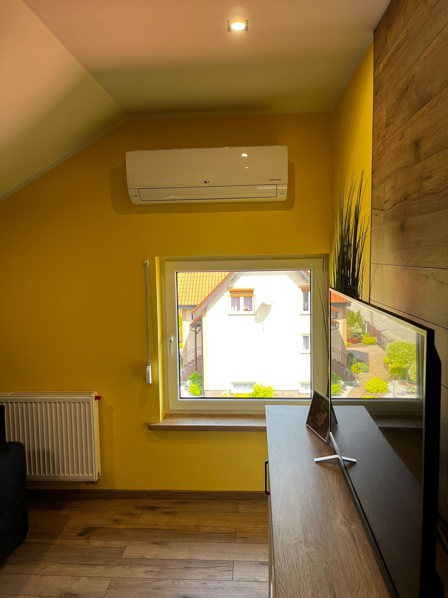 Biały klimatyzator LG zamontowany na żółtej ścianie w pokoju z oknem, widok na dom z ogrodem.