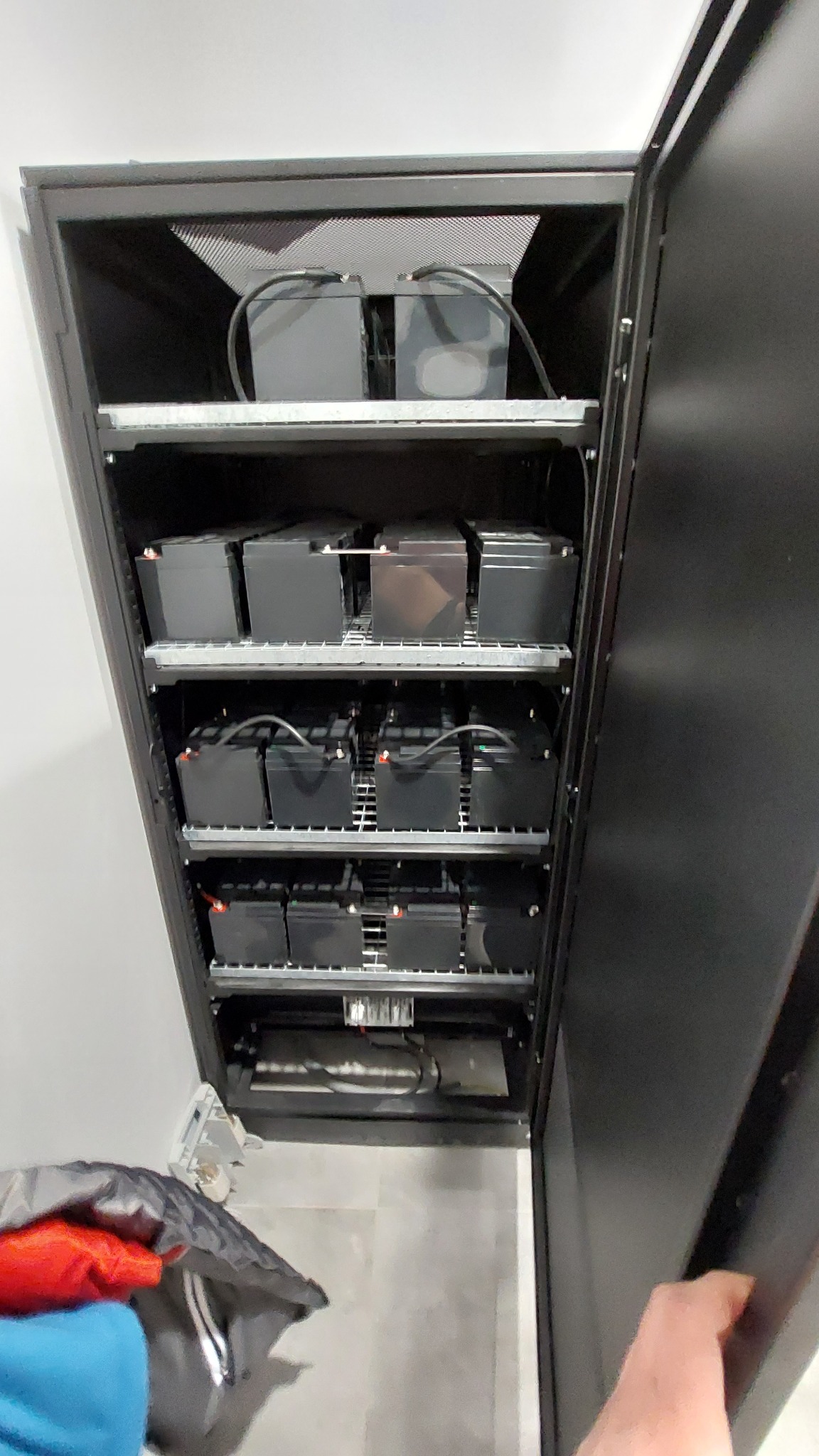 Szafa rack z zainstalowanymi akumulatorami, widoczne kable i metalowa konstrukcja, częściowo otwarte drzwi szafy.