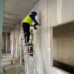 Pracownik w kasku i kamizelce na drabinie, montuje płytę na ścianie w surowym wnętrzu budynku, widoczne przewody elektryczne i elementy konstrukcyjne.