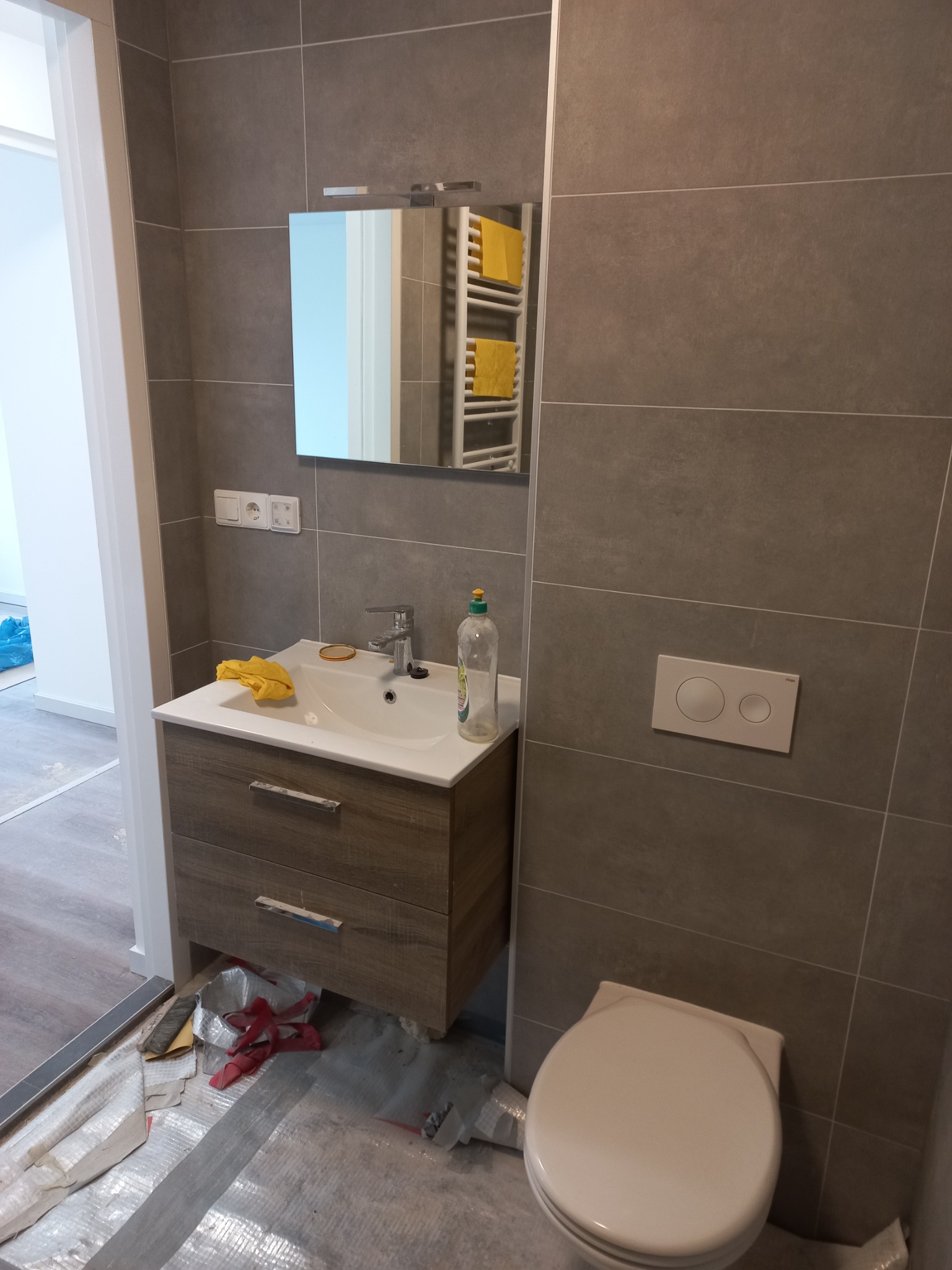 Nowoczesna łazienka w trakcie wykańczania: szare płytki, umywalka z drewnianą szafką, lustro z grzejnikiem drabinkowym i fragment toalety. Podłoga zabezpieczona folią, widoczne materiały budowlane.