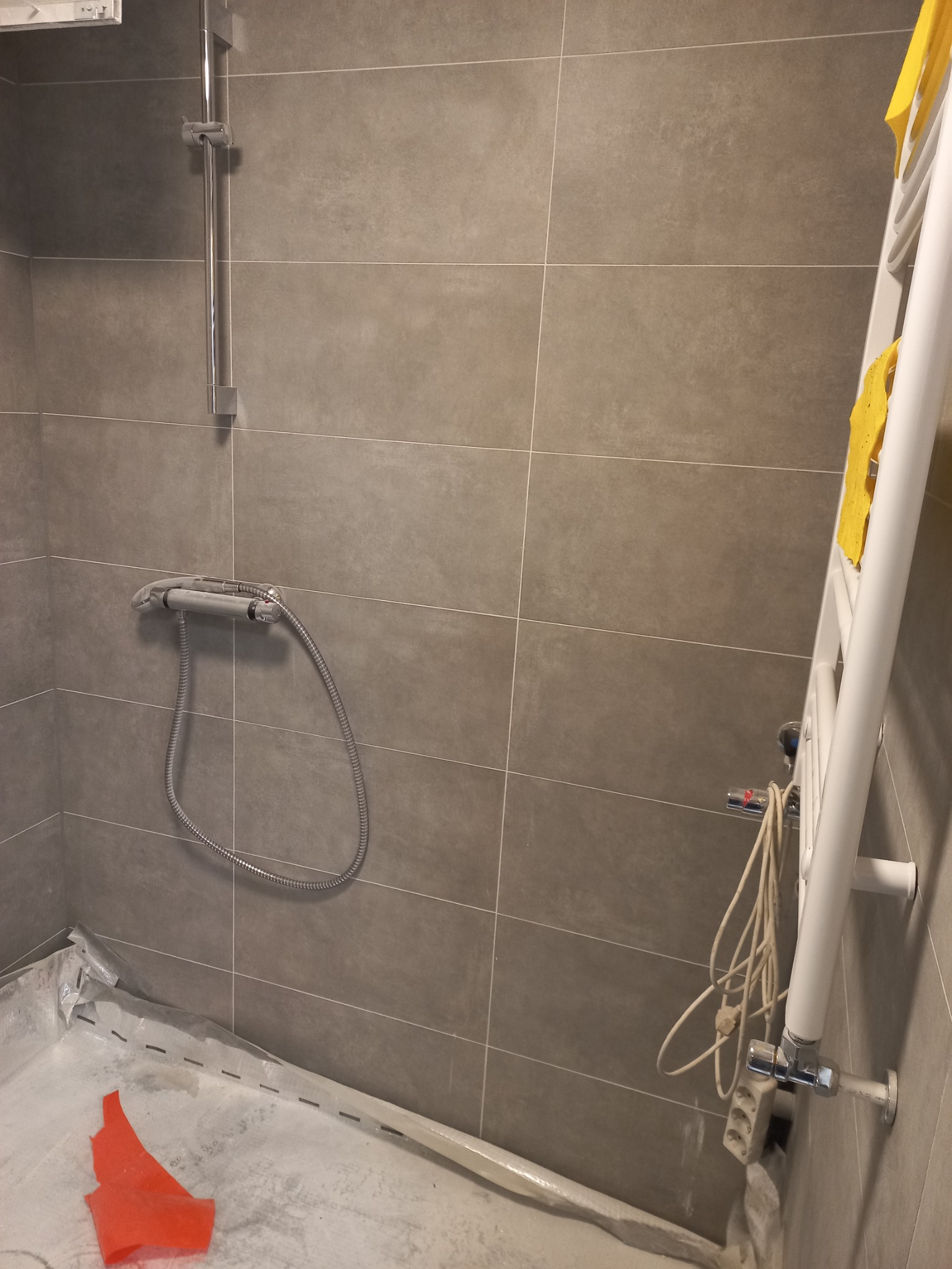 Remont łazienki: szare płytki na ścianach, chromowana bateria prysznicowa, zabezpieczona folią podłoga i grzejnik drabinkowy z żółtą szmatką. Widoczne narzędzia i przedłużacz.