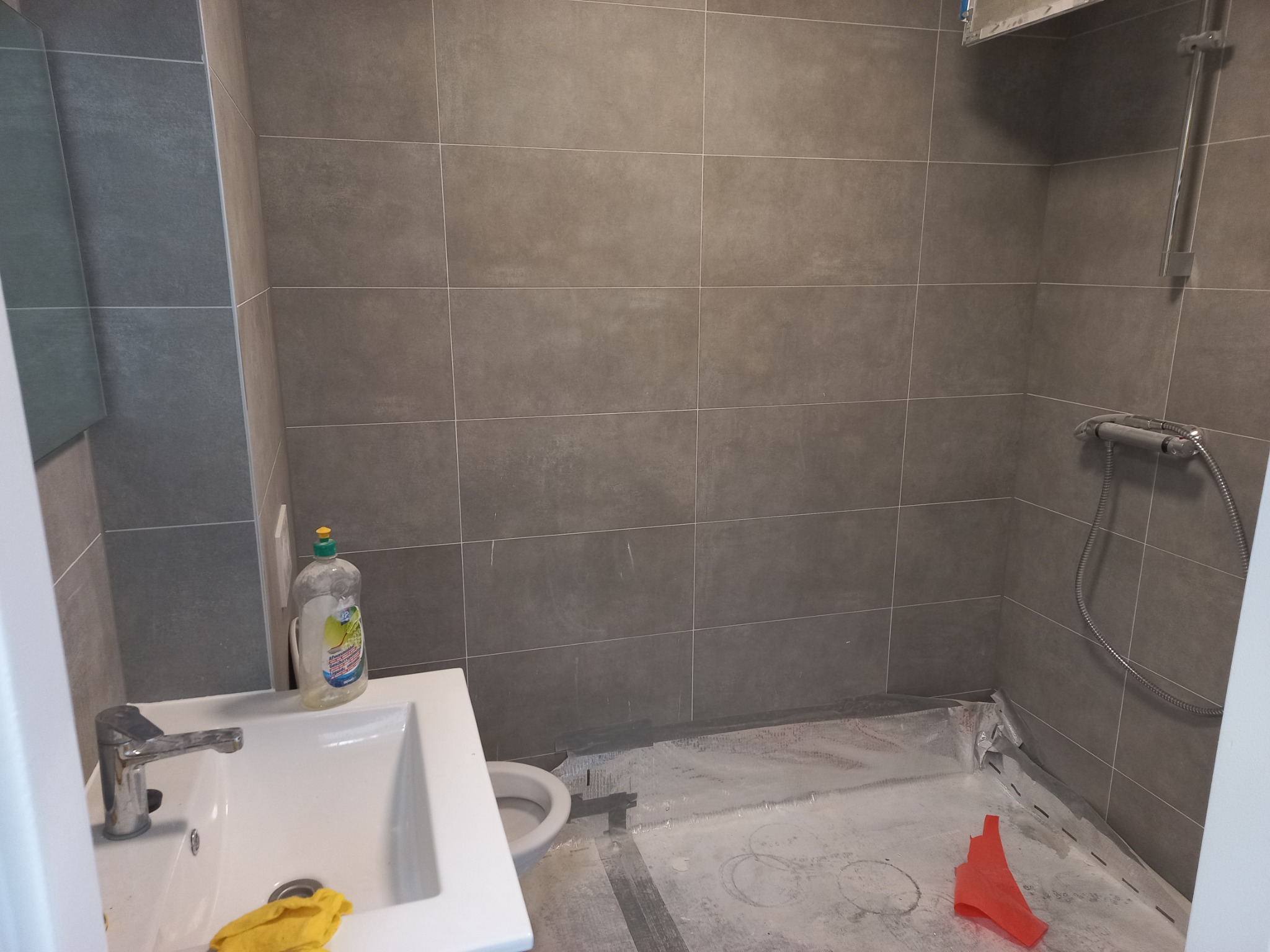 Łazienka w trakcie remontu z szarymi płytkami, zabezpieczona folią podłoga, widoczna umywalka z płynem do naczyń i czerwony element na podłodze.