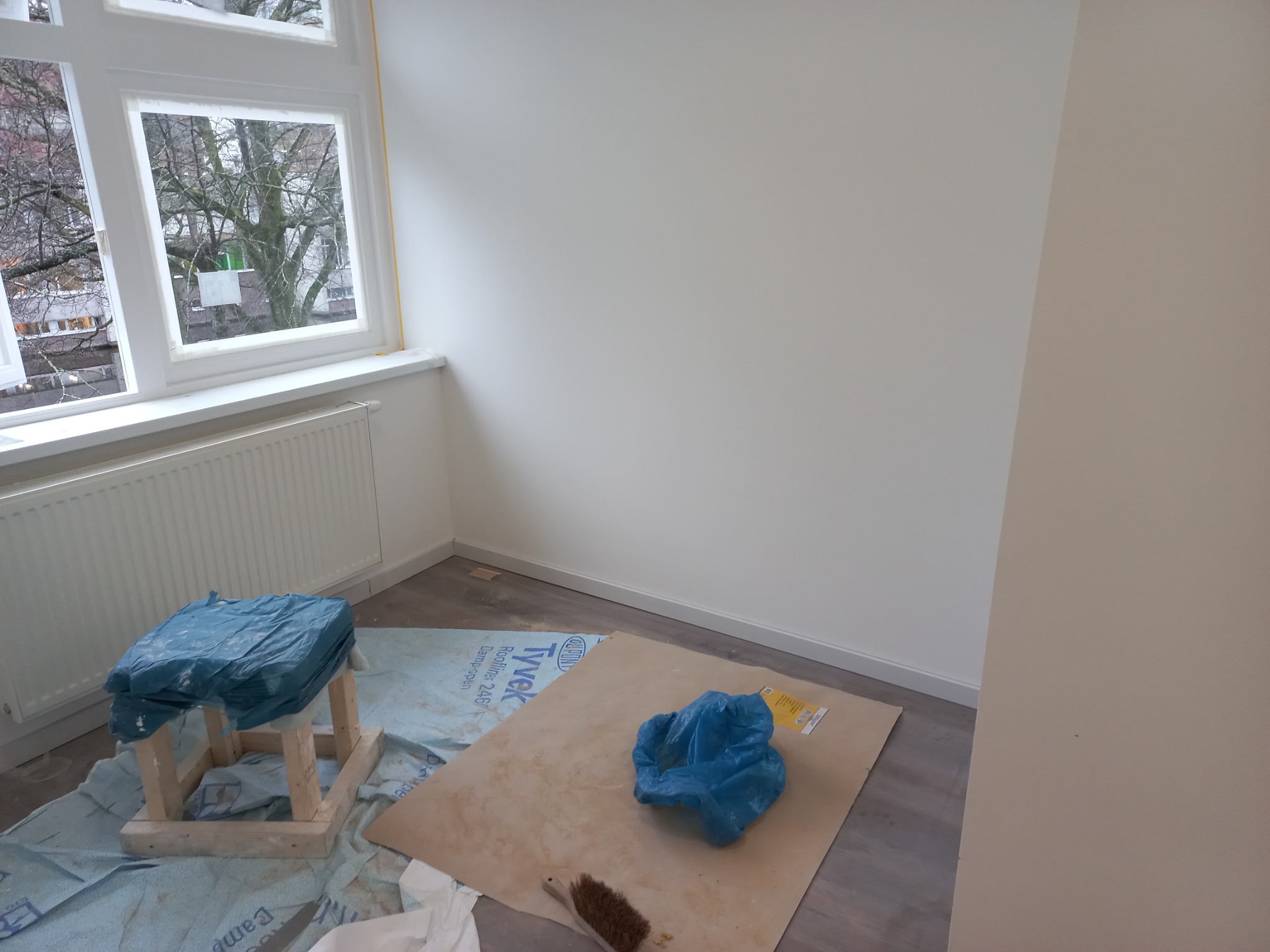 Pokój w trakcie malowania: zabezpieczone okno taśmą malarską, grzejnik, podłoga i meble przykryte folią ochronną, widoczna szczotka do odkurzania.
