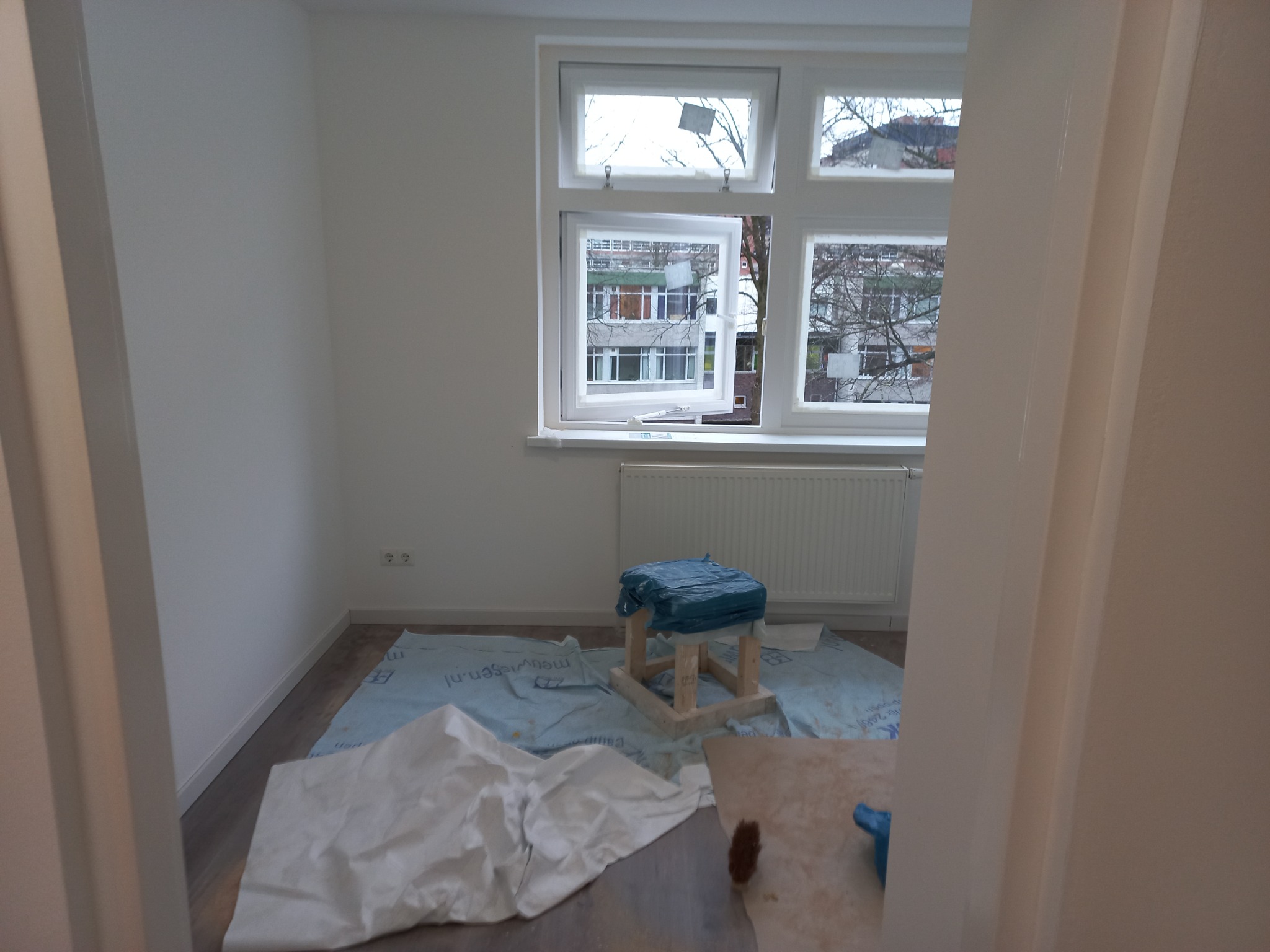 Pokój w trakcie przygotowań do malowania: zabezpieczona podłoga, okno z taśmą malarską, grzejnik i krzesło owinięte folią, widok zza framugi drzwi.
