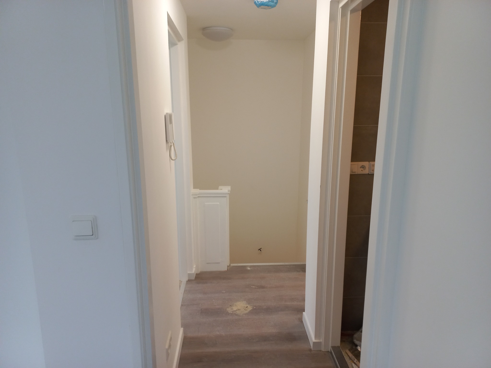 Korytarz w trakcie wykańczania, widoczne nowe panele podłogowe z plamą kleju, białe ściany i drzwi, w tle fragment łazienki z szarymi płytkami.