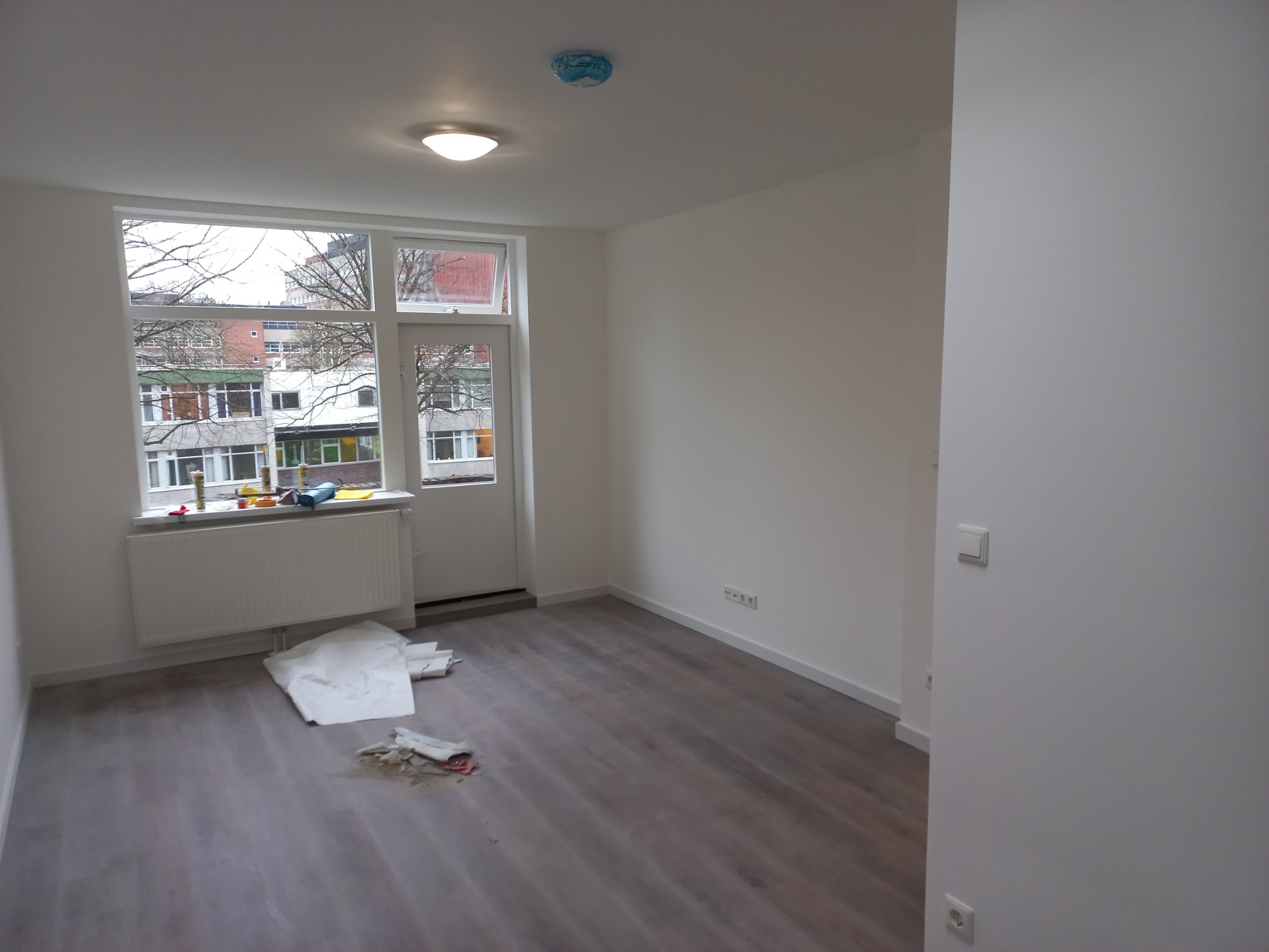 Pusty pokój po remoncie z nową podłogą i świeżo pomalowanymi ścianami na biało, zabezpieczony czujnik dymu na suficie, widok z okna na budynki.