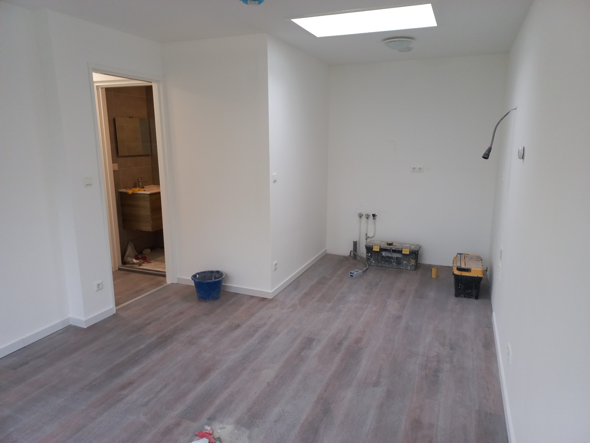 Pusty pokój po remoncie z panelami podłogowymi, widoczne narzędzia, wiadro i otwarte drzwi do łazienki z umywalką