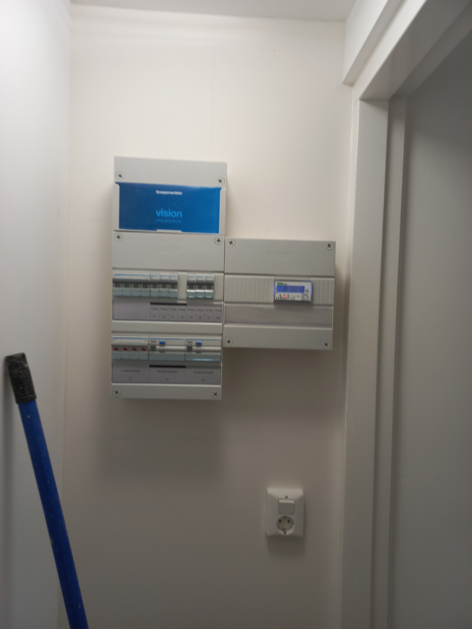 Rozdzielnica elektryczna Hager i Vision zamontowana na białej ścianie, obok widoczny fragment niebieskiego uchwytu pędzla malarskiego i gniazdko elektryczne.