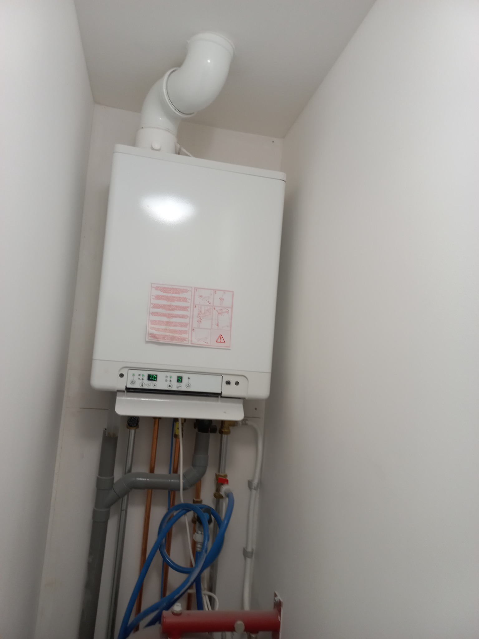 Biały, wiszący piec gazowy z podłączonymi rurami w jasnym pomieszczeniu, widoczne elementy instalacji grzewczej i wentylacyjnej.