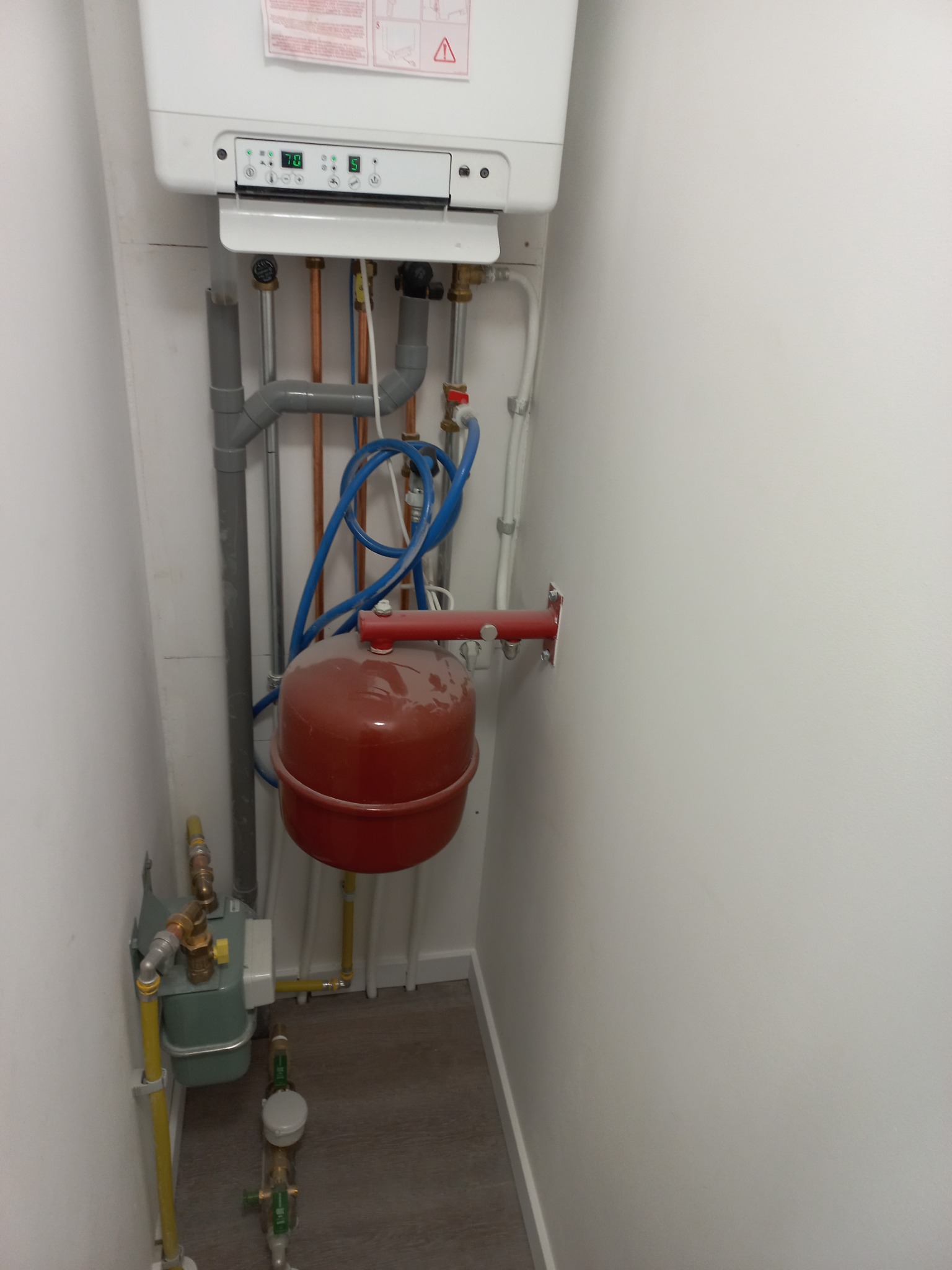 Zamontowany piec gazowy z podłączeniami hydraulicznymi i zbiornikiem wyrównawczym w małej wnęce z szarymi podłogami.