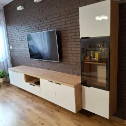 Nowoczesny zestaw mebli RTV z witryną na tle ściany z cegły. Białe fronty, drewniany blat, czarne uchwyty. W witrynie szkło i oświetlenie.