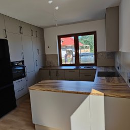 Jasnoszara kuchnia z drewnianym blatem i czarną lodówką. Widok na okno z widokiem na dom z czerwoną dachówką. Nowoczesny design, minimalistyczne uchwyty.