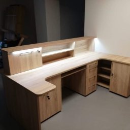 Jasna, narożna lada recepcyjna z oświetleniem LED, wykonana z drewna w odcieniach beżu. Widoczne szafki, półki i przestrzeń na komputer. Nowoczesny design.