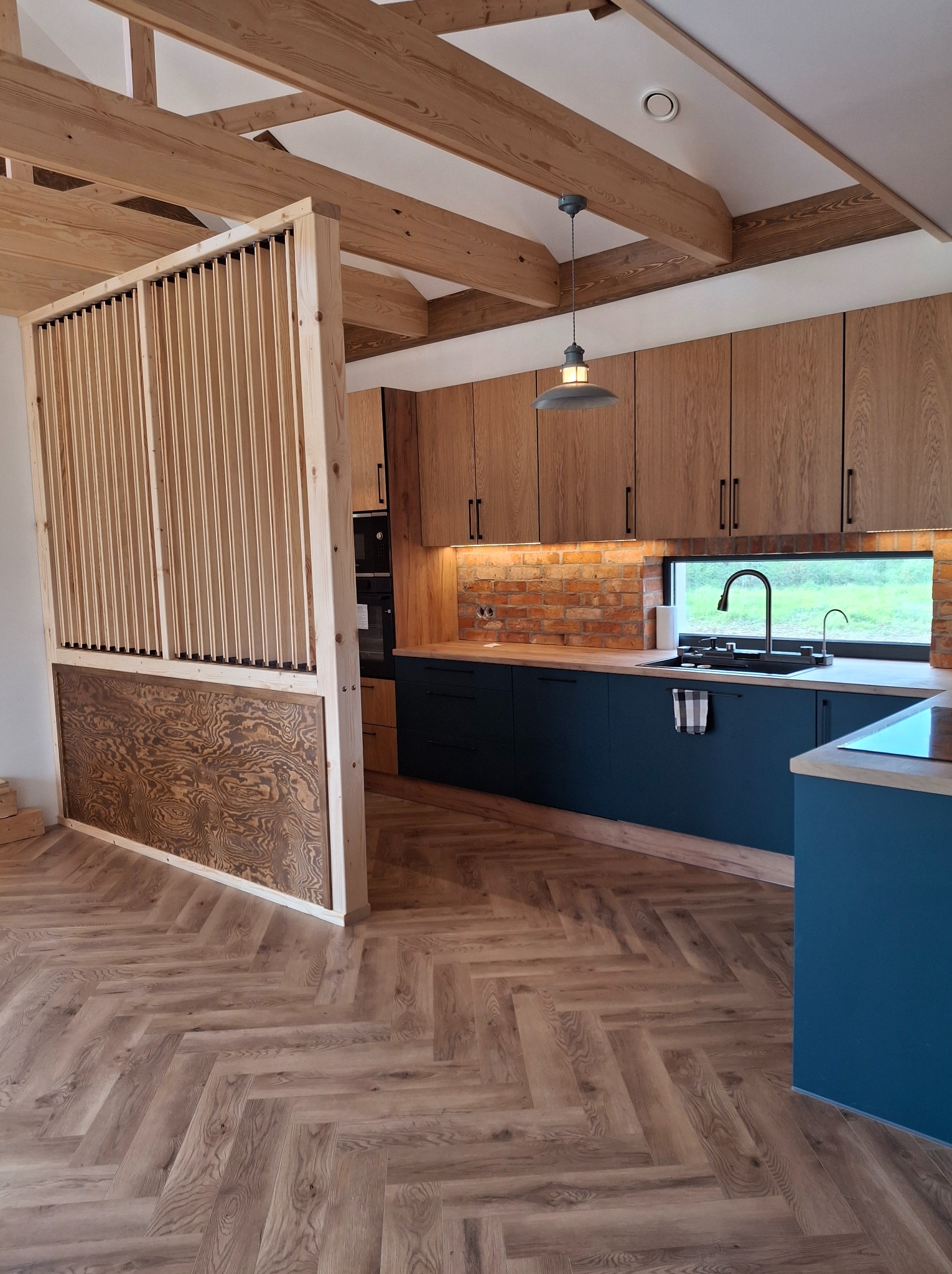 Nowoczesna kuchnia z drewnianymi szafkami, ciemnoniebieskimi dolnymi szafkami, ceglaną ścianą nad blatem i drewnianą ażurową ścianką działową. Parkiet w jodełkę na podłodze.