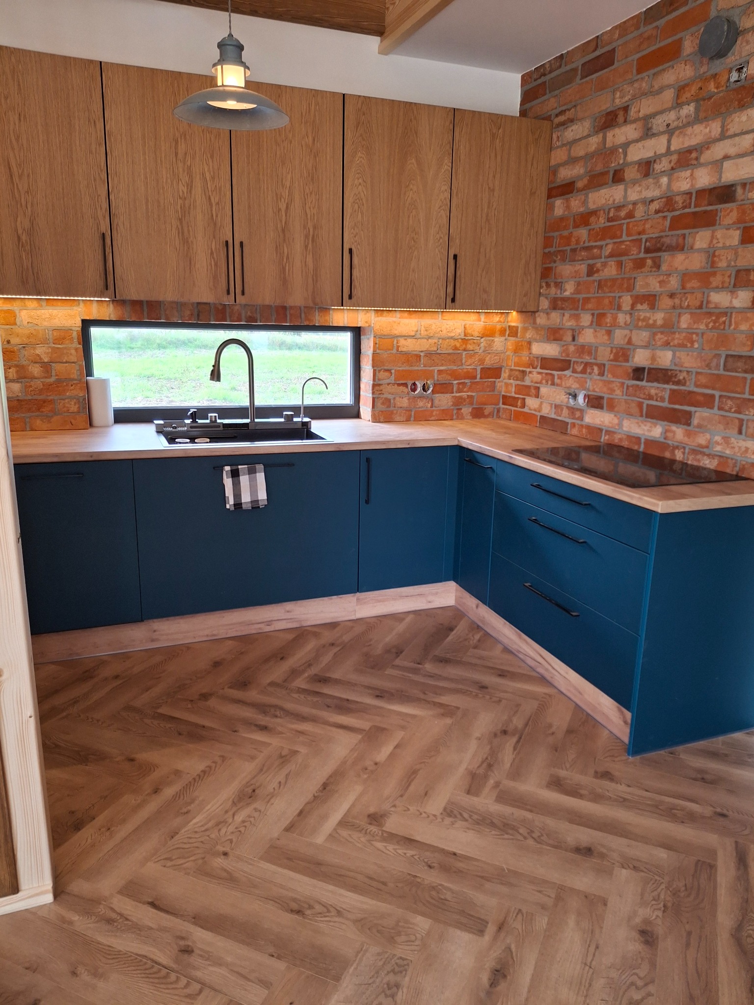 Nowoczesna kuchnia z drewnianymi szafkami górnymi, niebieskimi szafkami dolnymi i ceglaną ścianą, oświetlona lampą wiszącą i listwą LED, z drewnianą podłogą ułożoną w jodełkę.